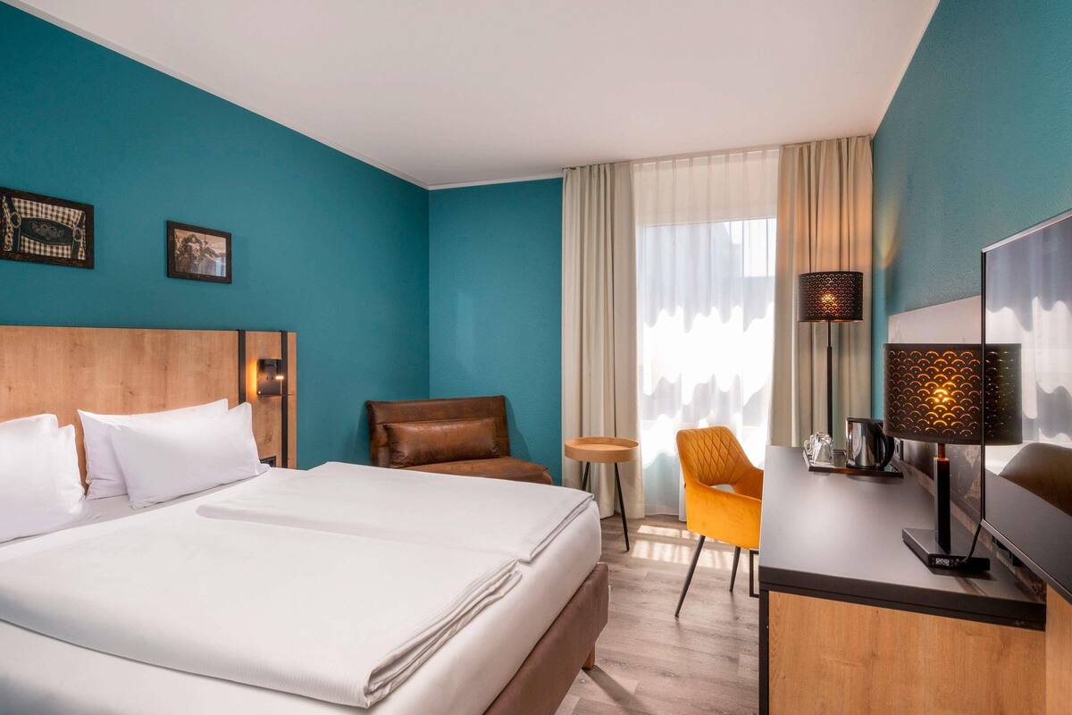 Quarto de hotel com cama, escrivaninha, cadeira e janela com cortinas.