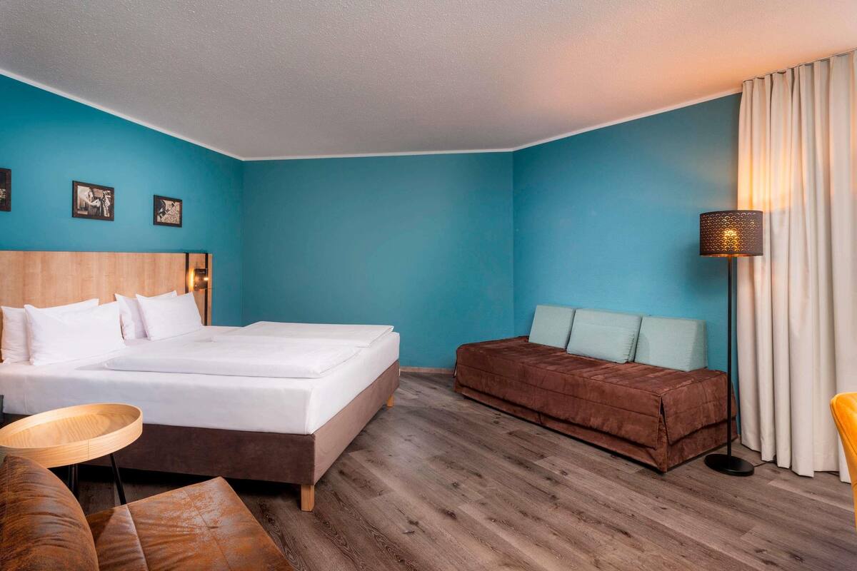 A hotel room with a bed, a sofa, a small round table, and a floor lamp.