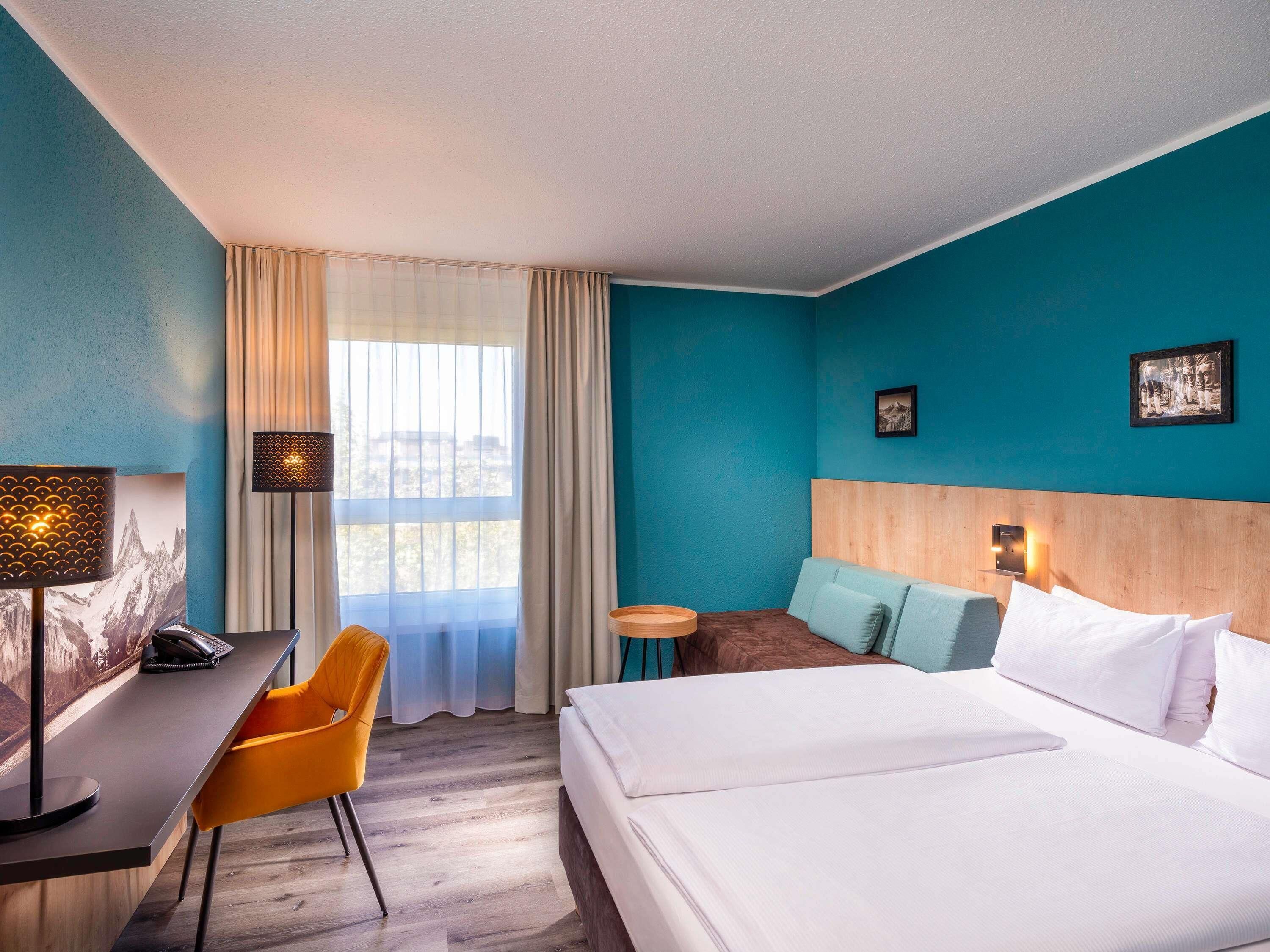 Une chambre d’hôtel avec un lit, un bureau, une chaise, une petite table et une fenêtre avec des rideaux.