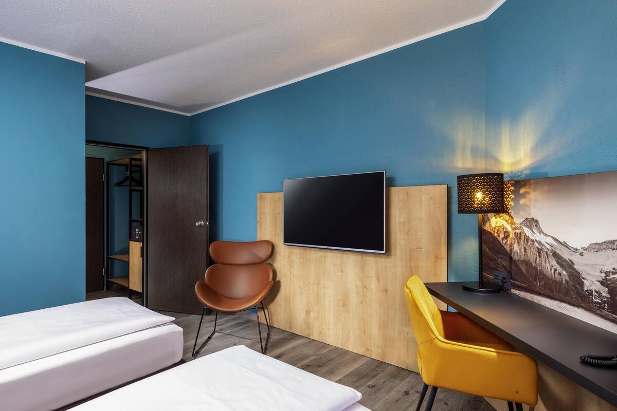 A hotel room with a bed, a desk with a TV, a chair, and a wall-mounted lamp.