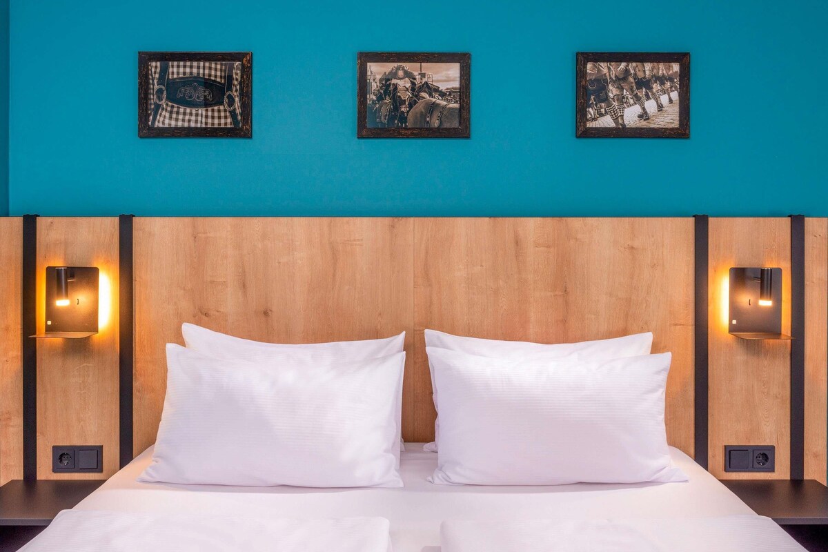Habitación de hotel con cabecera de madera, dos almohadas blancas y tres cuadros en la pared.