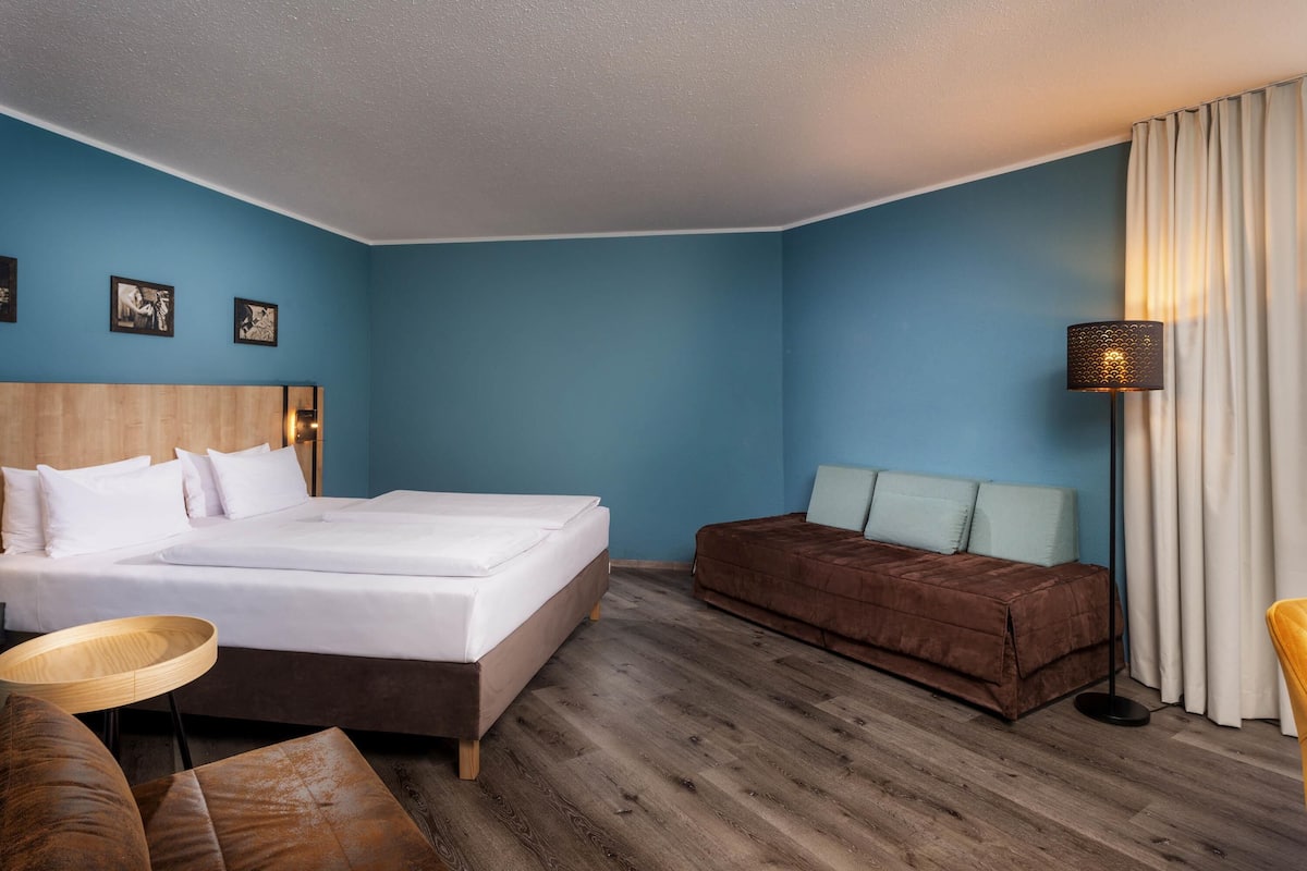 Quarto de hotel com cama, sofá, mesa de madeira redonda e abajur.
