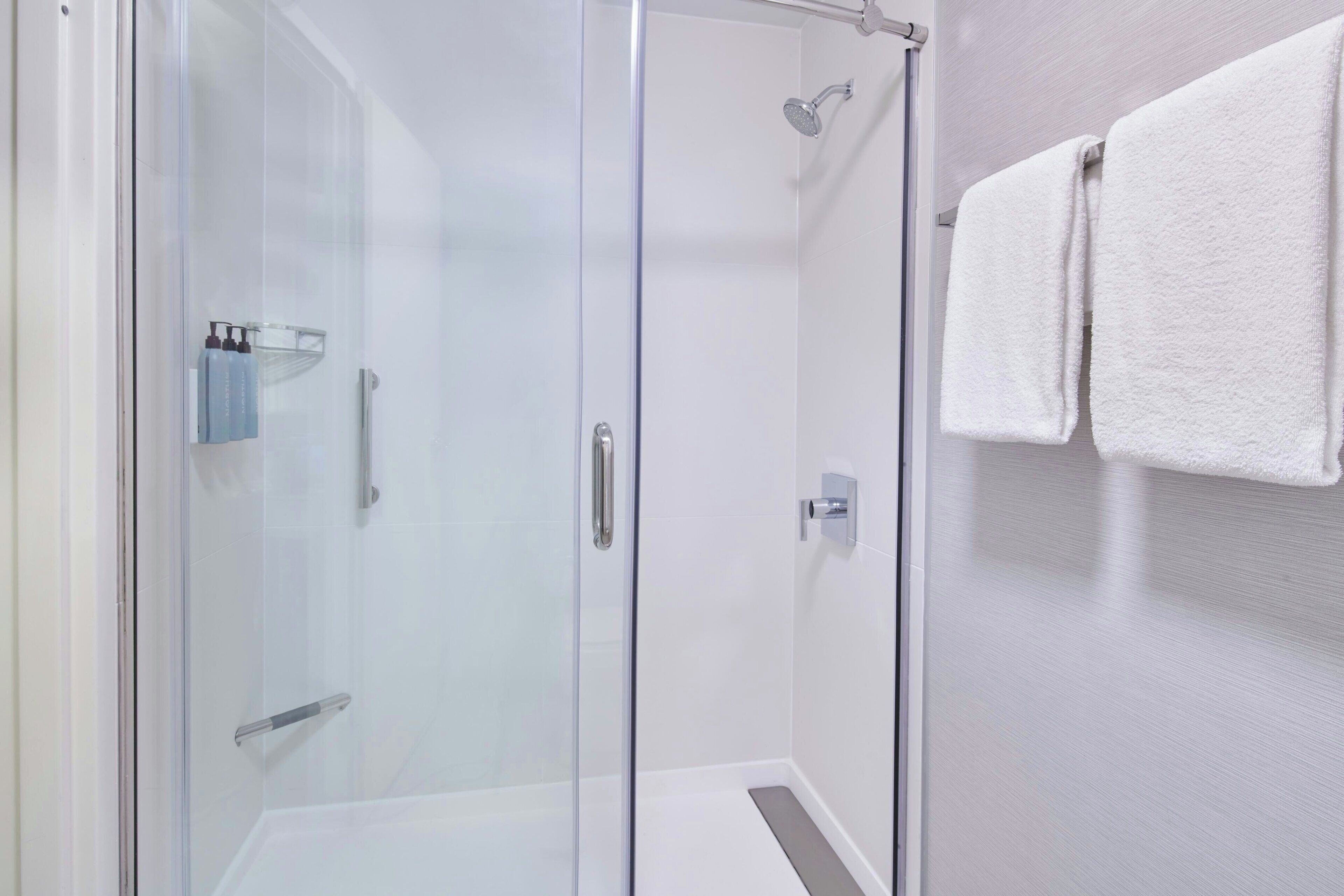 Box doccia in vetro con soffione a mano, ripiano in vetro con articoli da toilette e porta asciugamani con due asciugamani.