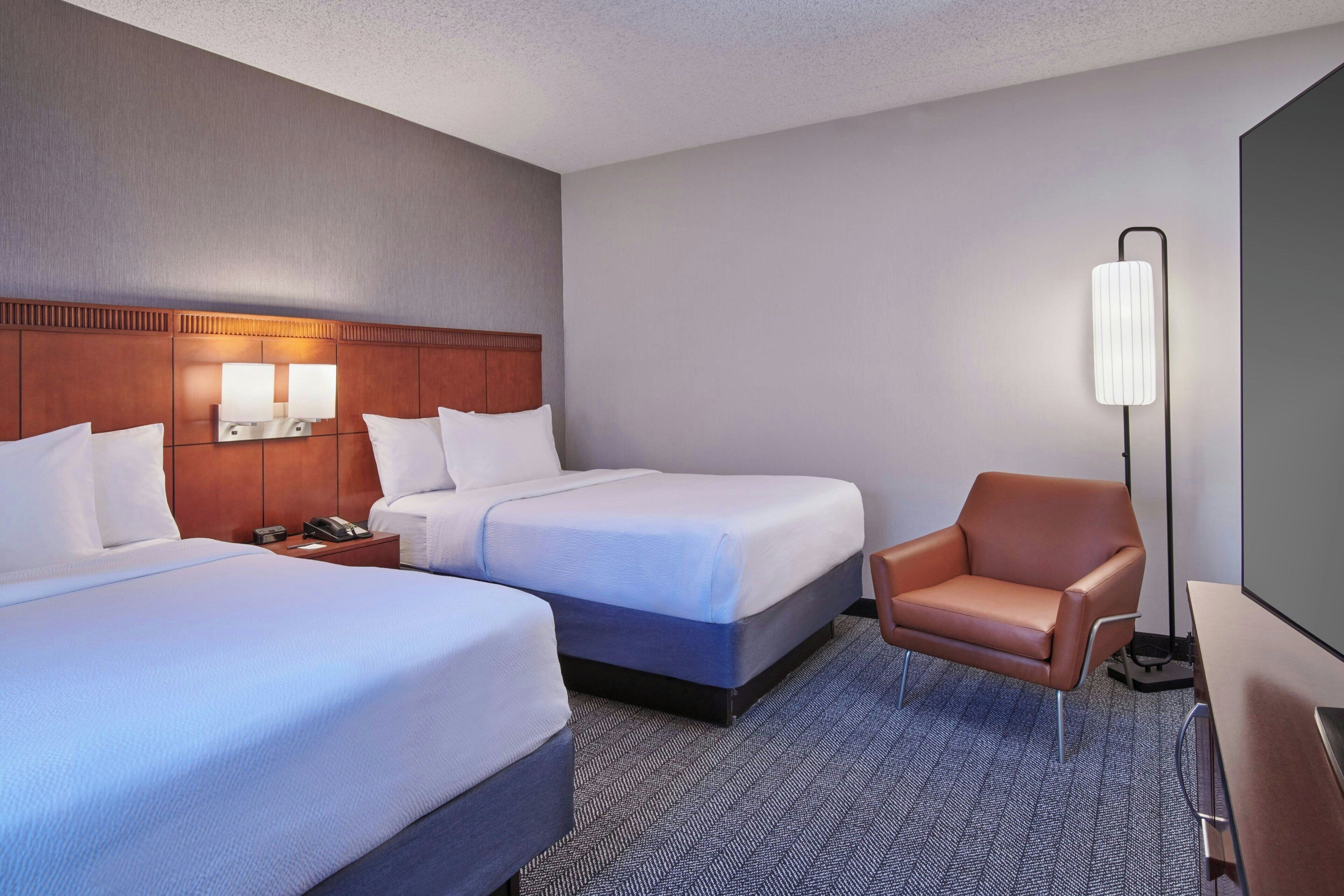 A hotel room with two beds, a desk, a chair, and a standing lamp.