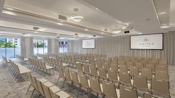 Ein Konferenzraum mit Reihen beiger Stühle, einem großen Bildschirm, der 'AMORA BRISBANE' zeigt, und Blick auf die Stadt durch große Fenster.