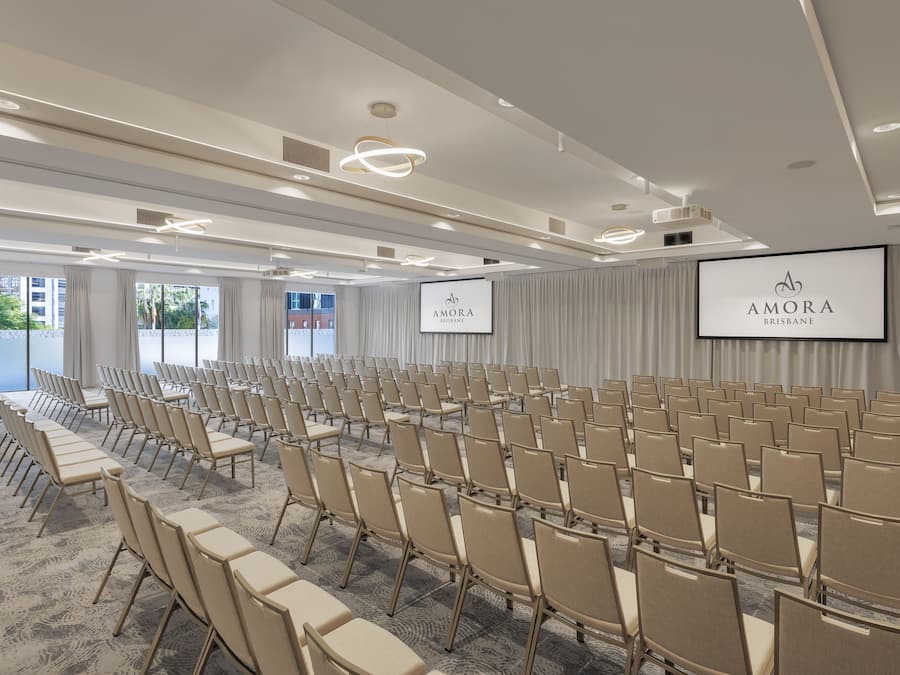 Ein Konferenzraum mit Reihen beiger Stühle, einem großen Bildschirm, der 'AMORA BRISBANE' zeigt, und Blick auf die Stadt durch große Fenster.