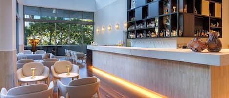 A modern bar area with a wooden floor, seating with round tables, and a bar counter with shelves of bottles.