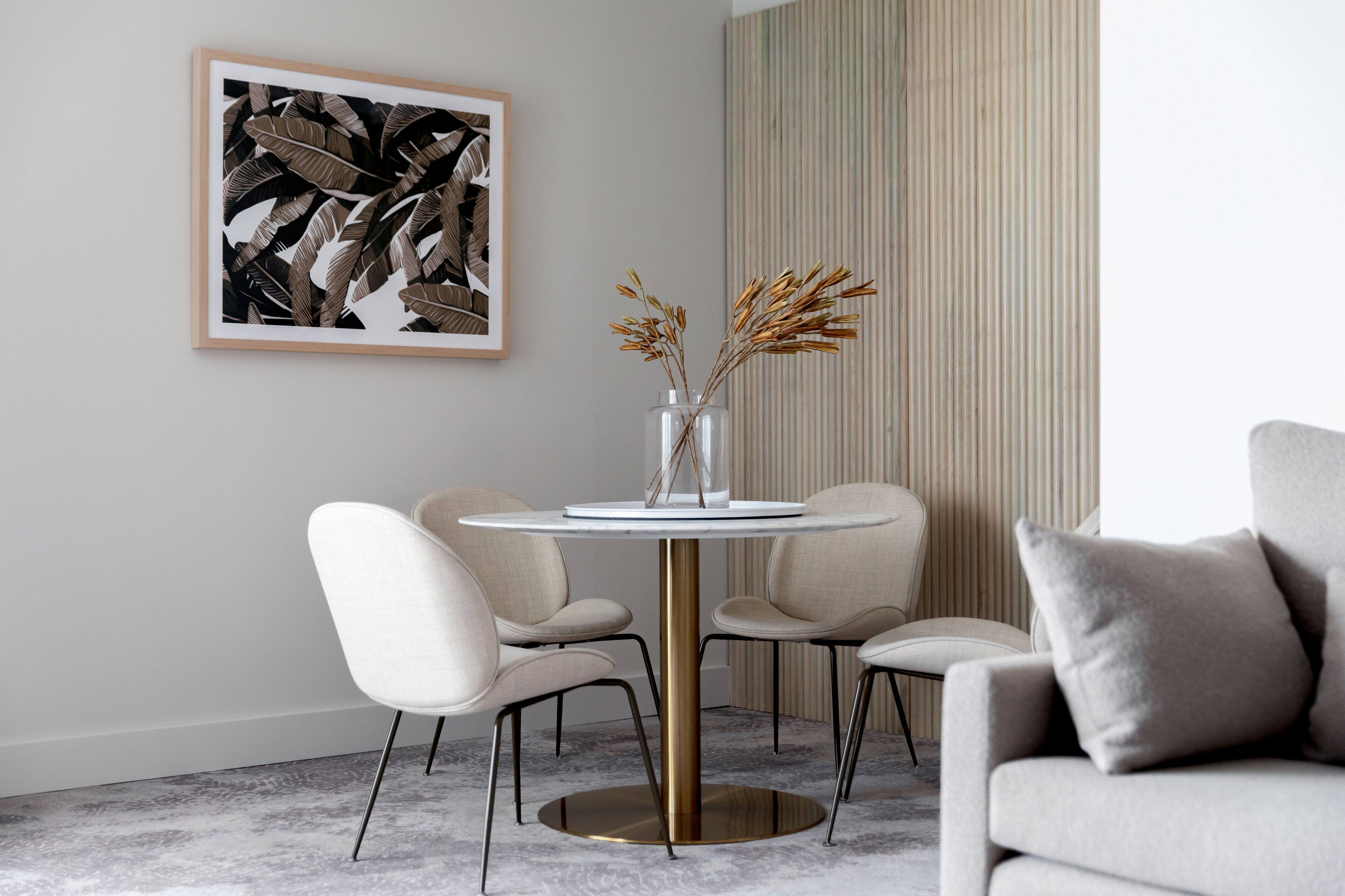 Un comedor moderno con una mesa redonda, cuatro sillas, un jarrón con plantas secas y una obra de arte enmarcada en la pared.