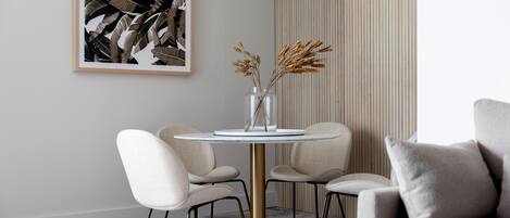 A modern dining area with a round table, four chairs, a vase with dried plants, and a framed artwork on the wall.