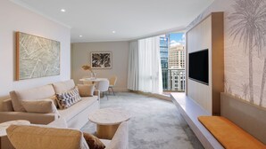 A modern hotel room with a sofa, armchair, and a small table. There is a TV mounted on the wall and a window with a view of buildings. - Amora Hotel Brisbane (Spring Hill)