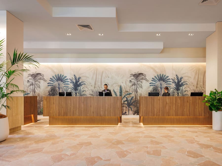 A reception area with two wooden reception desks, a large wall mural of palm trees, and potted plants.
