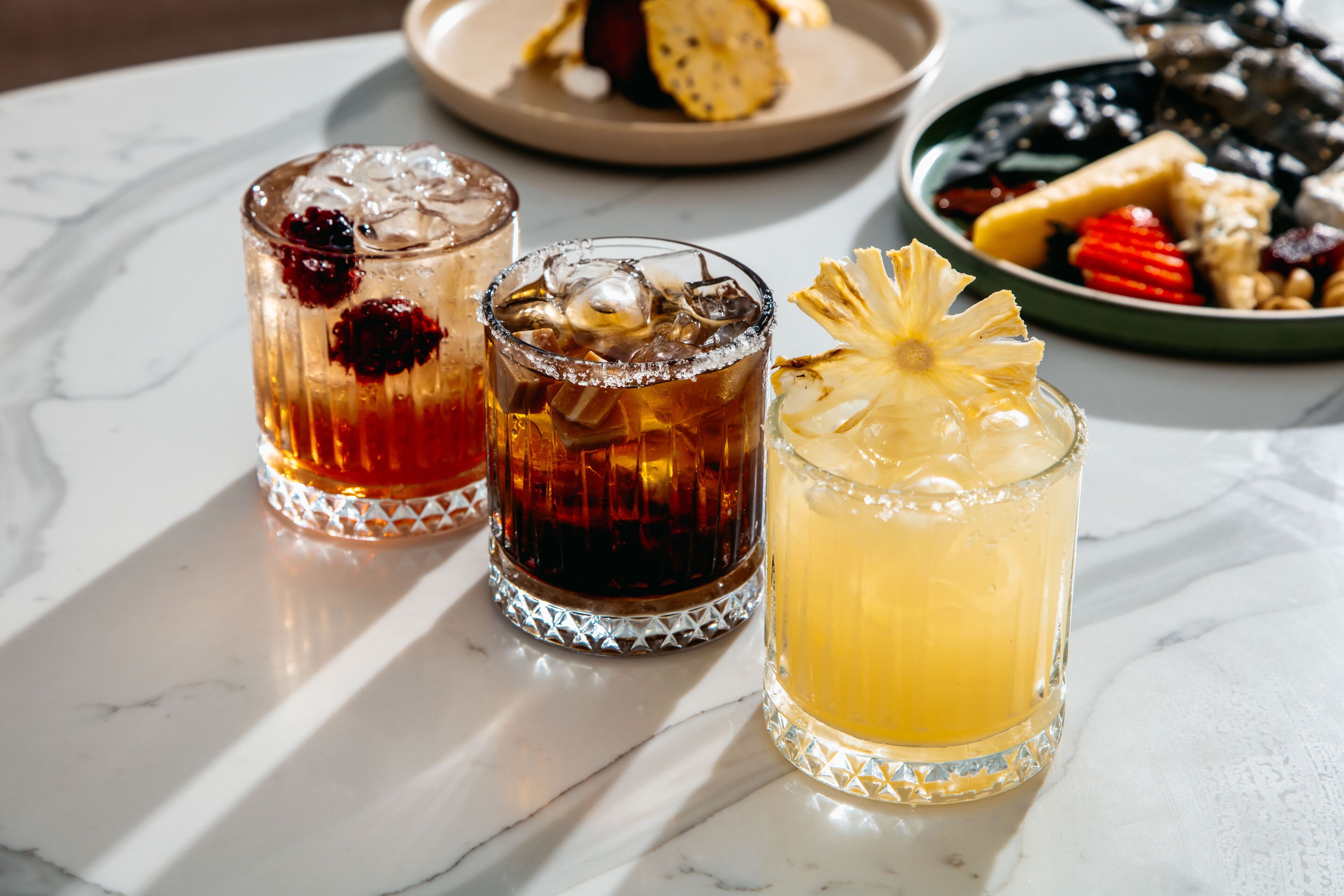 Three glasses of beverages on a marble table with a plate of assorted fruits and a bowl of nuts in the background.