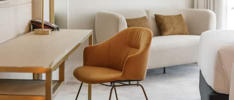 A hotel room with a wooden desk, a modern orange chair, a sofa with cushions, and a framed picture on the wall.
