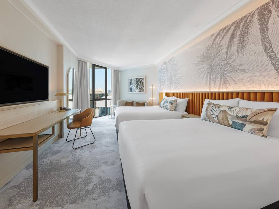 A hotel room with a large flat-screen TV, a desk with a chair, two beds with white linens, a large window with curtains, and a wall mural of palm trees.