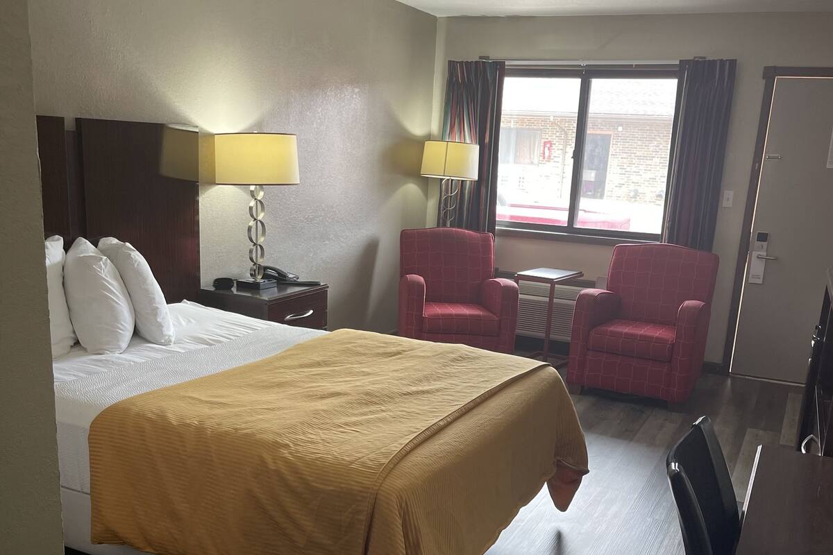 A hotel room with a bed, two chairs, a small table, and a window with curtains.