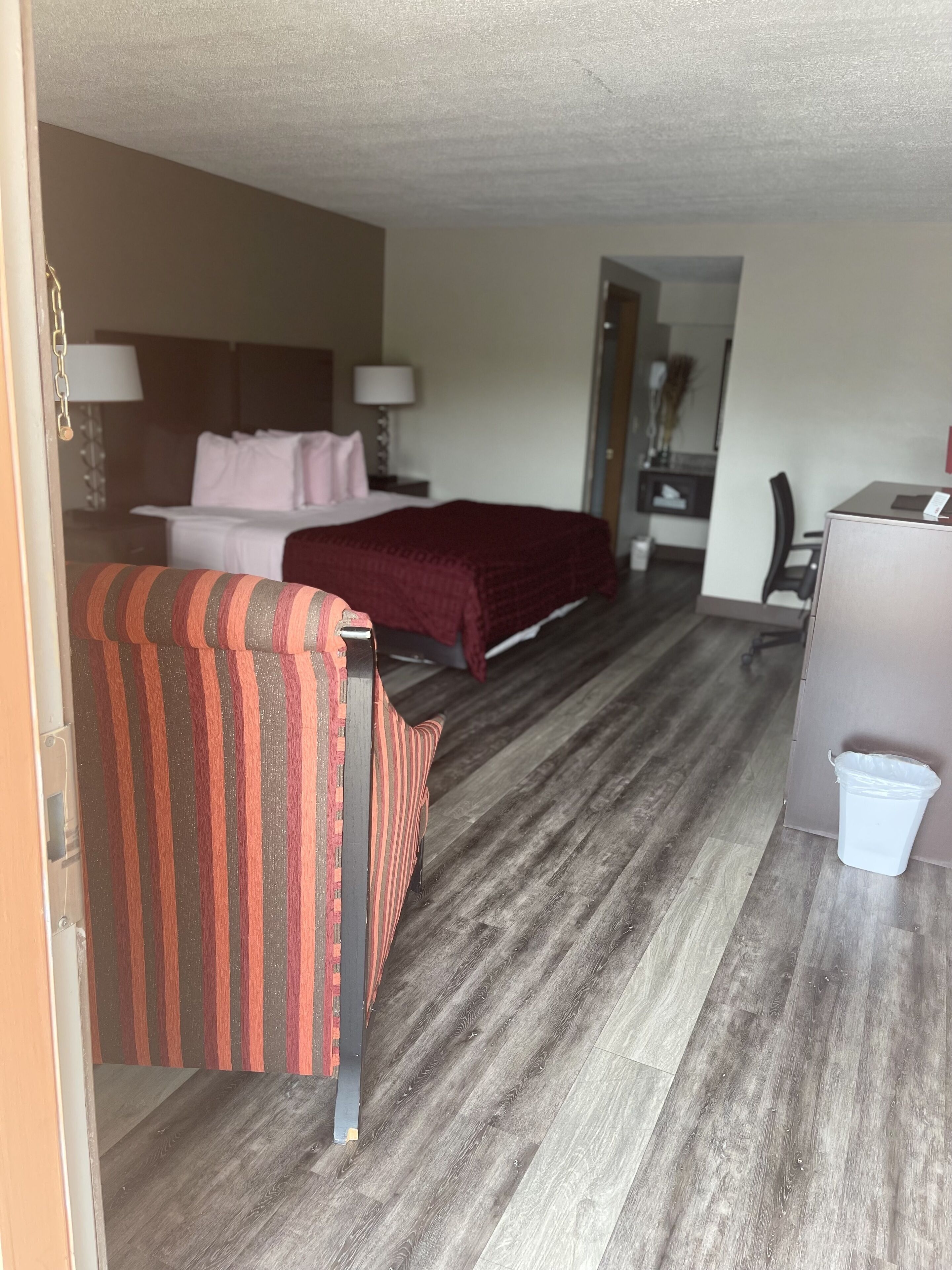 A hotel room with a bed, a striped chair, a desk, and a trash bin.