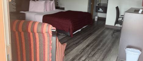 A hotel room with a bed, a striped chair, a desk, and a trash bin.