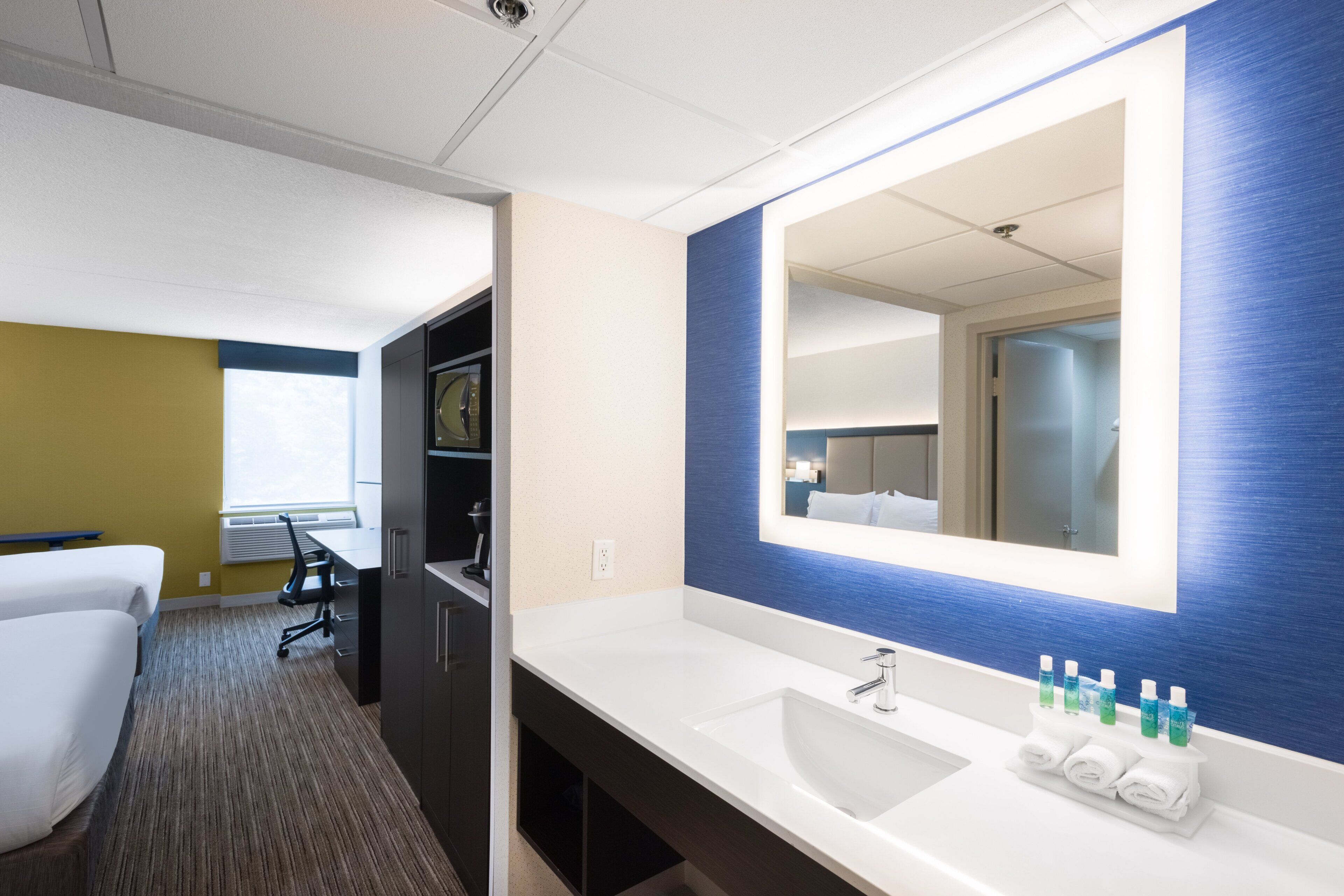 A hotel room with a large mirror, a white sink, and a small table with toiletries.