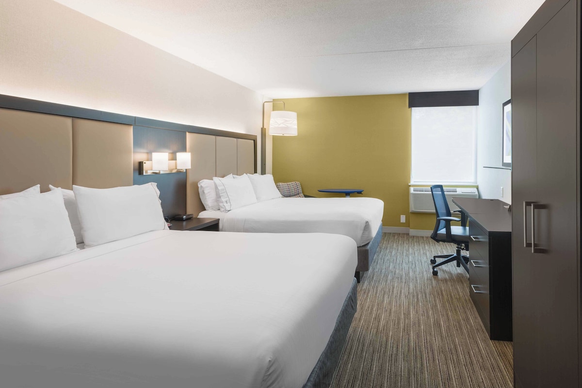A hotel room with two beds, a desk, a chair, a lamp, and a window.