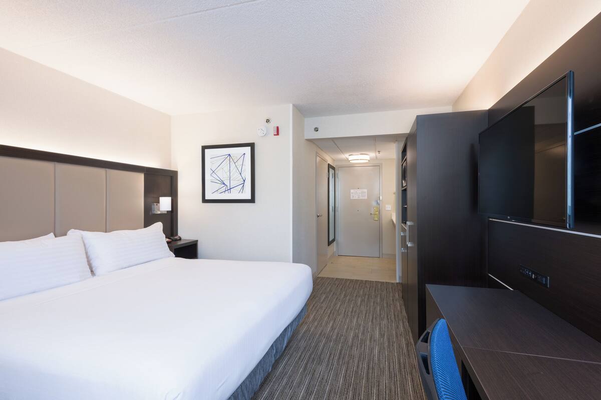 A hotel room with a large bed, a desk with a flat-screen TV, a framed picture on the wall, and a door leading to a hallway.