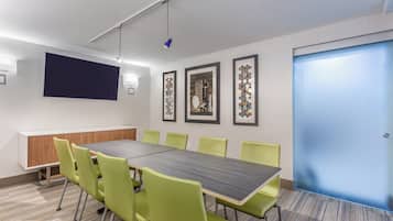 A conference room with a long table, green chairs, a projector screen, and framed artwork on the walls.