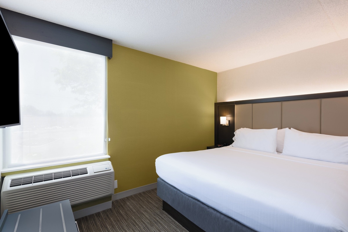A hotel room with a bed, a window, an air conditioning unit, and a wall-mounted light.