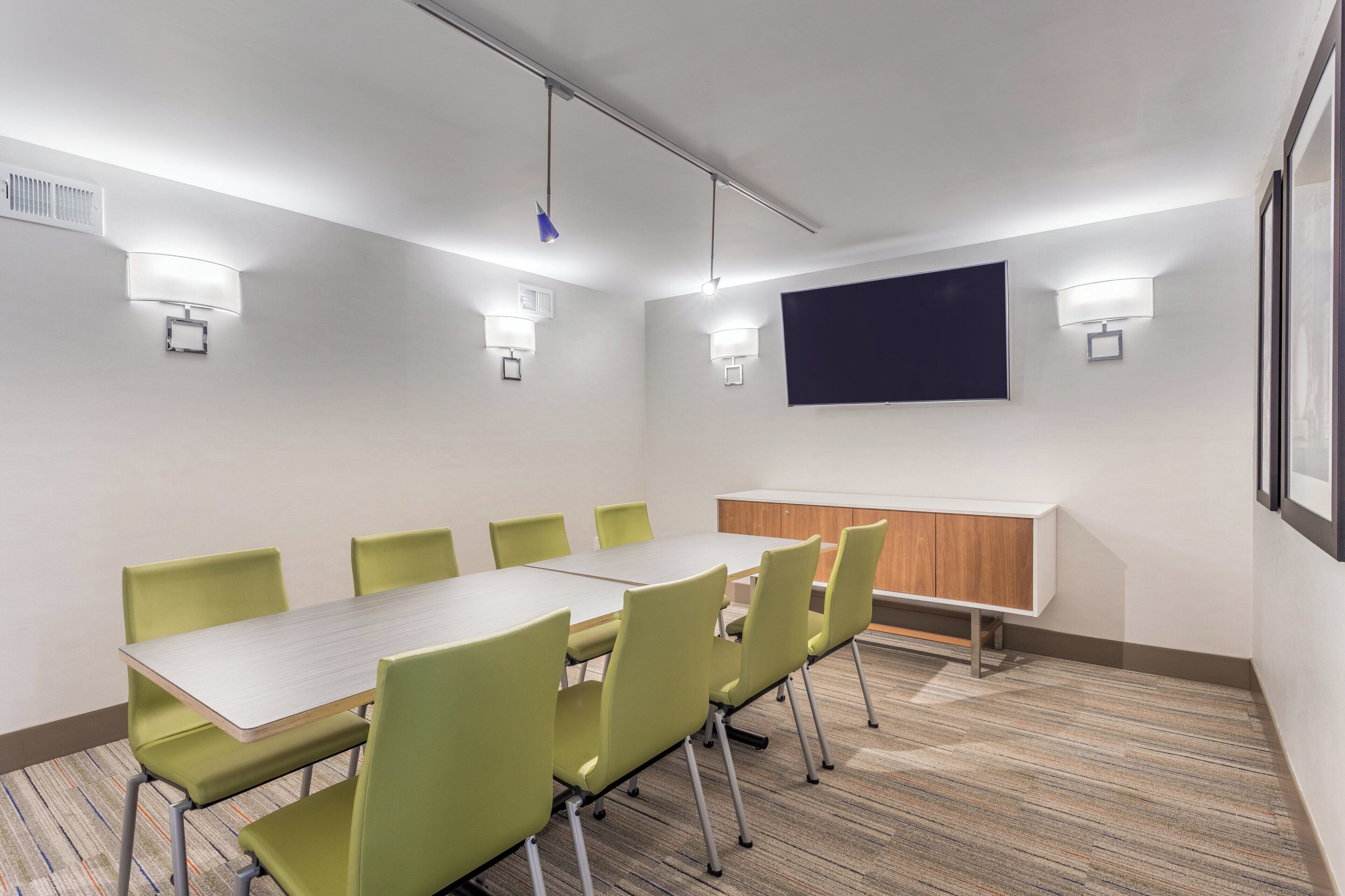 A conference room with a long table, green chairs, a whiteboard, and wall-mounted lights.