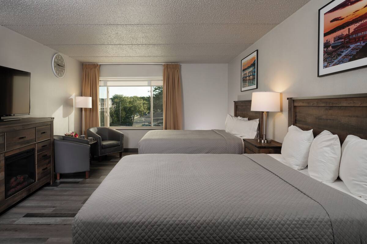 Une chambre d’hôtel avec deux lits, une télévision, un bureau avec une chaise, une cheminée et une grande fenêtre avec des rideaux.