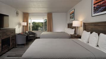 A hotel room with two beds, a TV, a desk with a chair, a fireplace, and a large window with curtains.