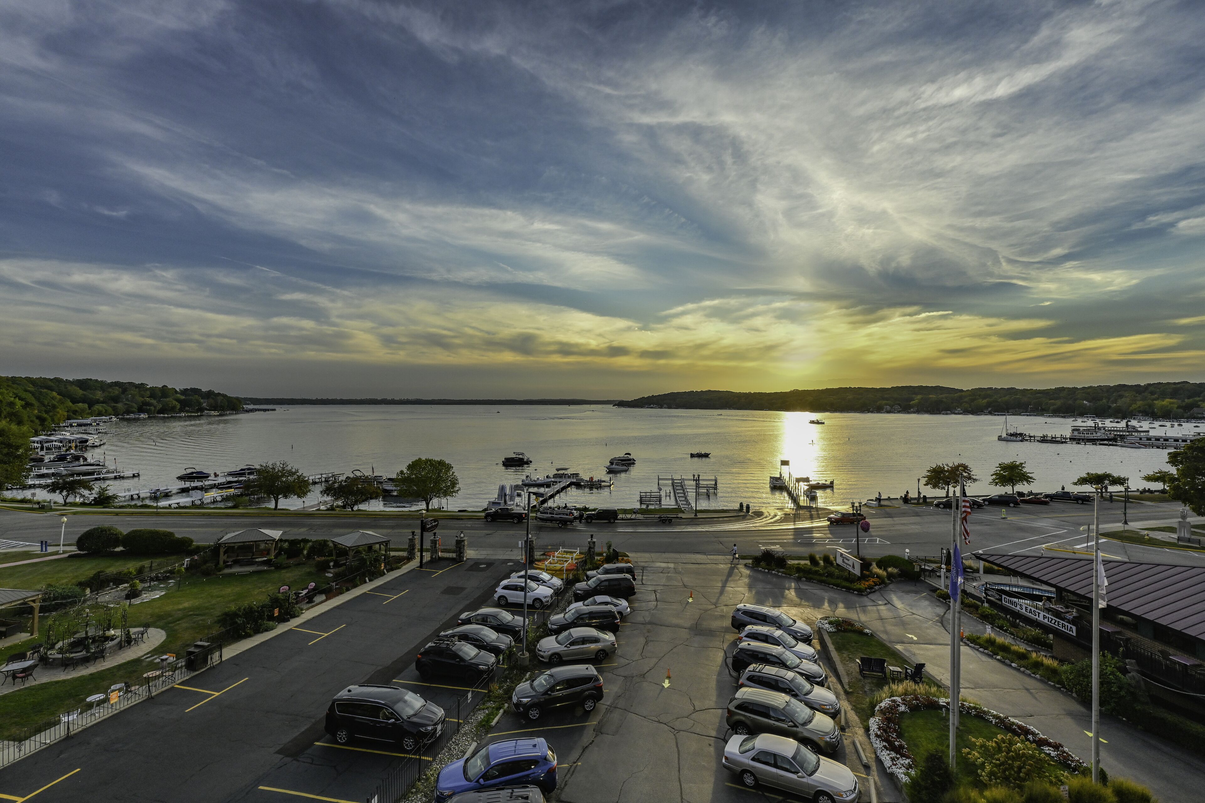 Un parking rempli de voitures, un port de plaisance avec des bateaux, et un coucher de soleil sur une étendue d’eau.