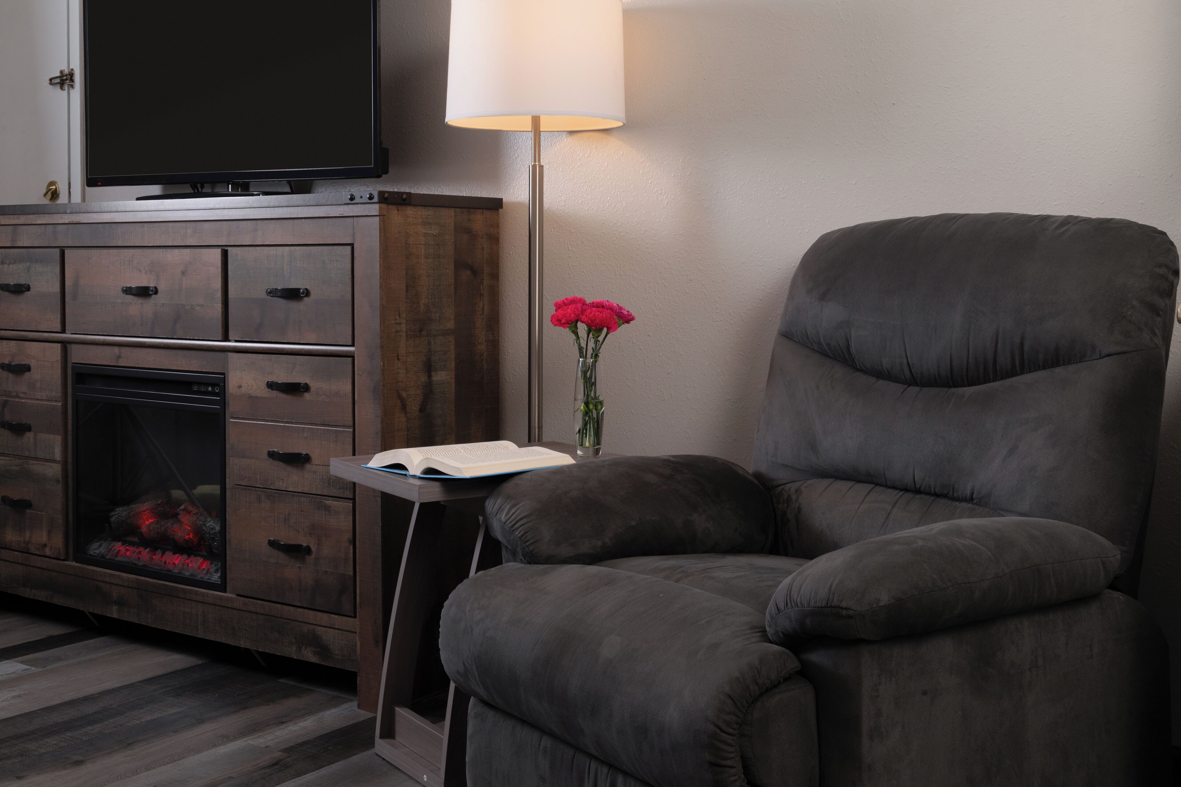 A modern living room with a dark grey armchair, a flat-screen TV, a wooden entertainment unit, a small side table with a book and a vase of red flowers.