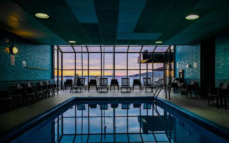 An indoor swimming pool with a view of the sunset through large windows, surrounded by lounge chairs.