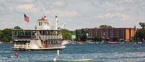 A riverboat with a prominent American flag, a lighthouse, and a building with a red roof in the background.