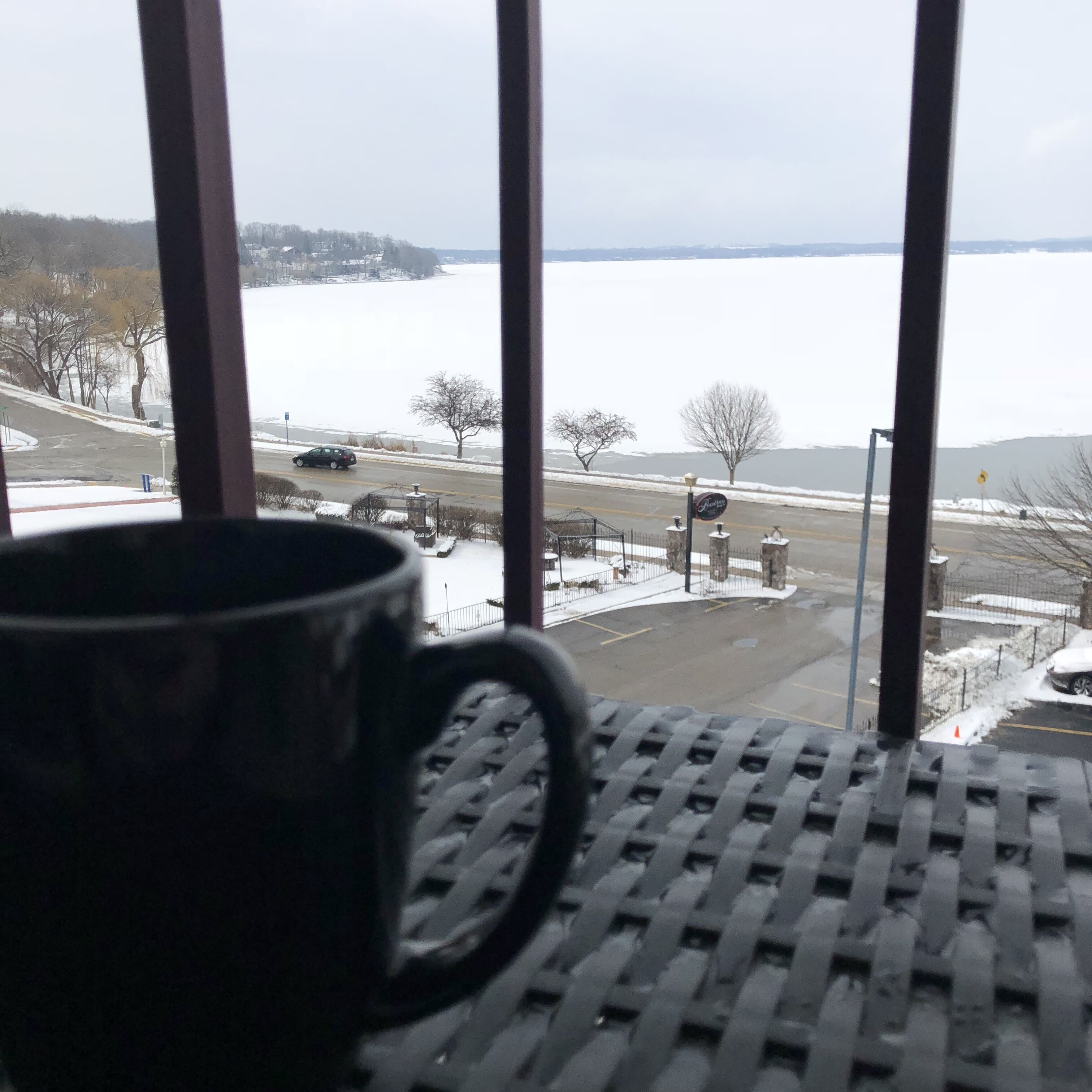 A view from a window with a black mug on a table, overlooking a snowy landscape with a road and a body of water.
