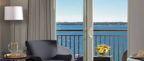 A hotel room with a bed, a chair, a table with a pitcher and glasses, a vase with flowers, and a view of the water.