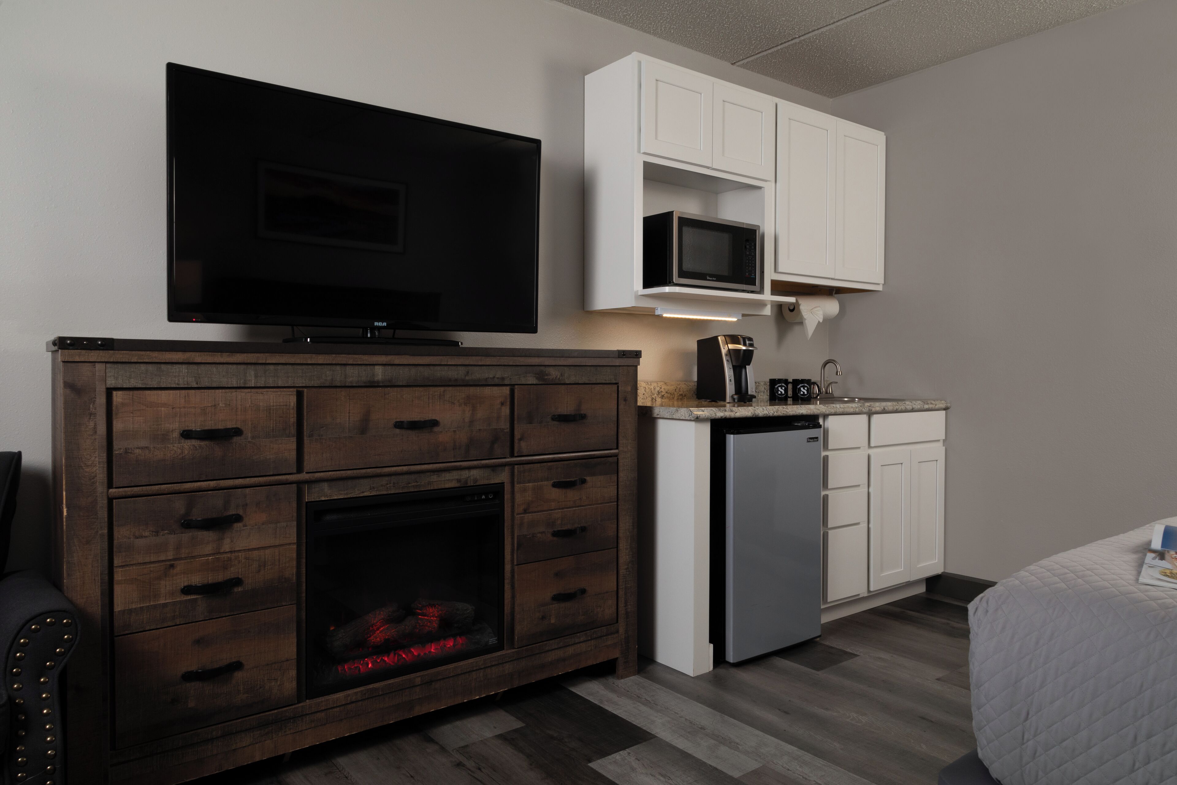 Una habitación compacta con una televisión de pantalla plana montada sobre un mueble bajo de madera con cajones. Un microondas se encuentra sobre una encimera blanca, y se aprecia un lavamanos pequeño.