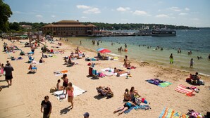 Une plage où des gens se prélassent au soleil et se baignent, une jetée avec des bateaux, et un bâtiment en arrière-plan.