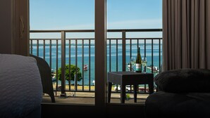 Une chambre d’hôtel avec un balcon donnant sur une étendue d’eau, une table avec une bouteille et un verre, et une vue sur des bateaux et un arbre.