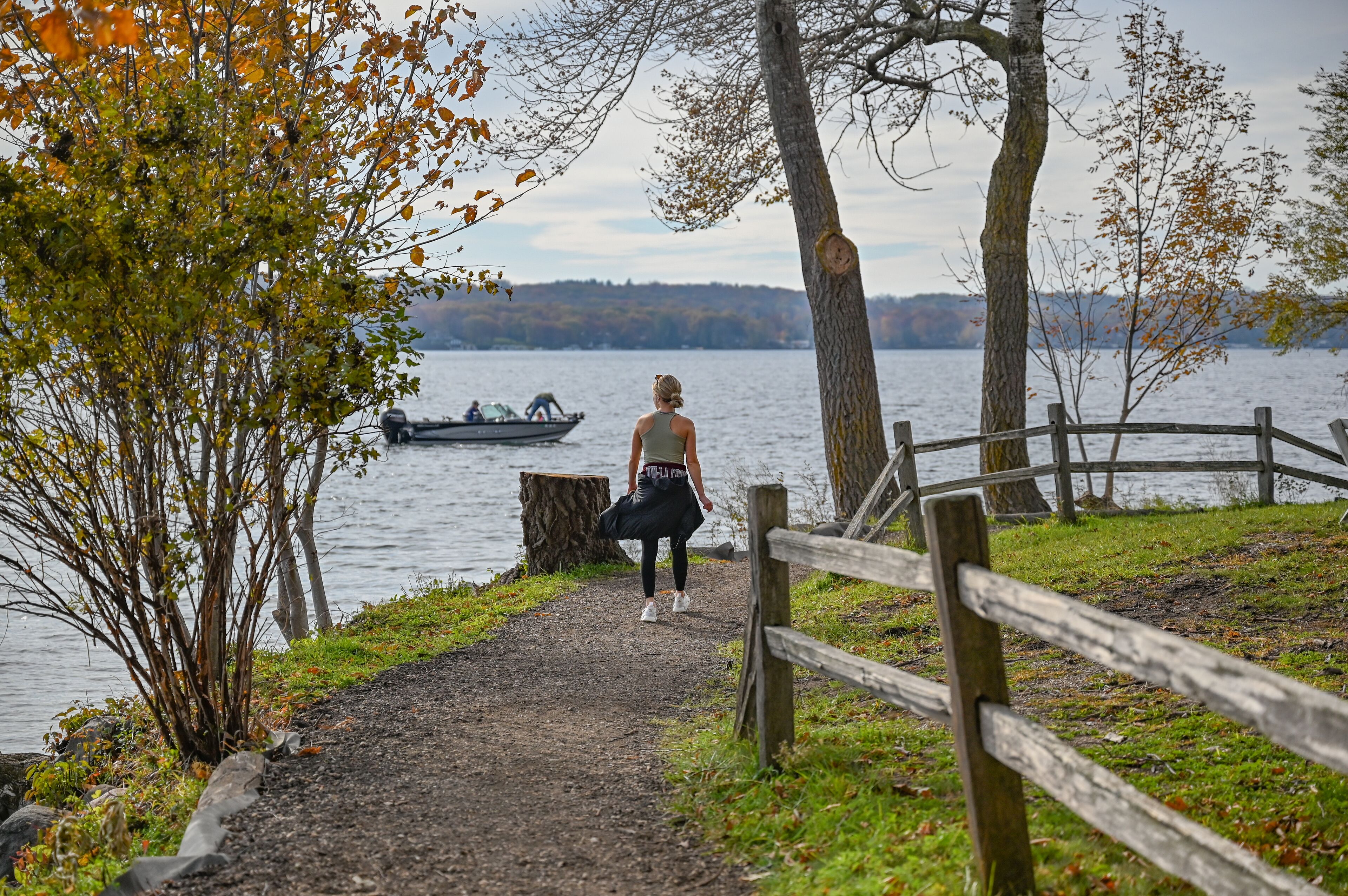Une personne marchant sur un sentier de terre au bord d’un lac, avec un bateau sur l’eau et des arbres en arrière-plan.
