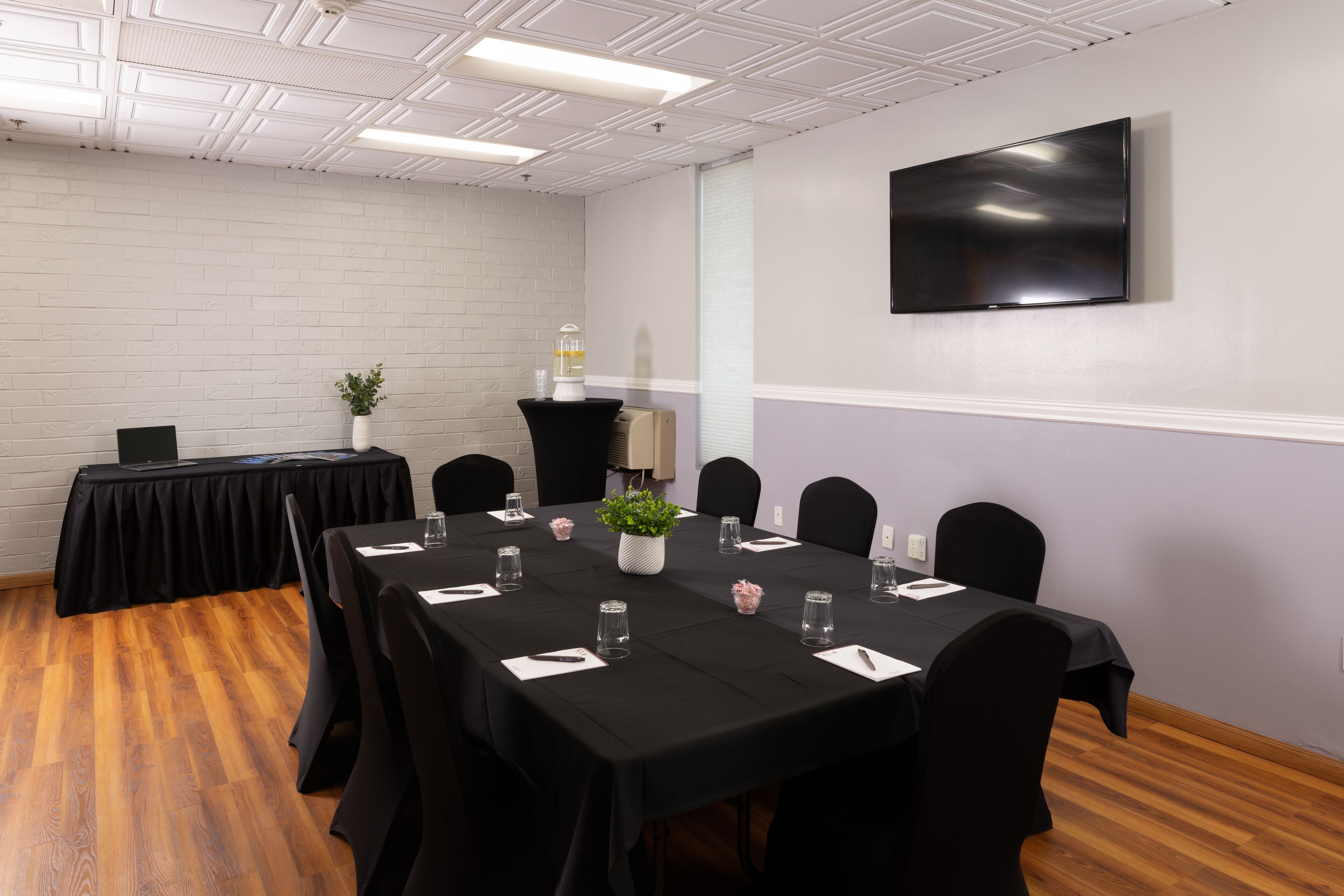 A conference room with a long table, black chairs, a white brick wall, a flat-screen TV, and a small plant.