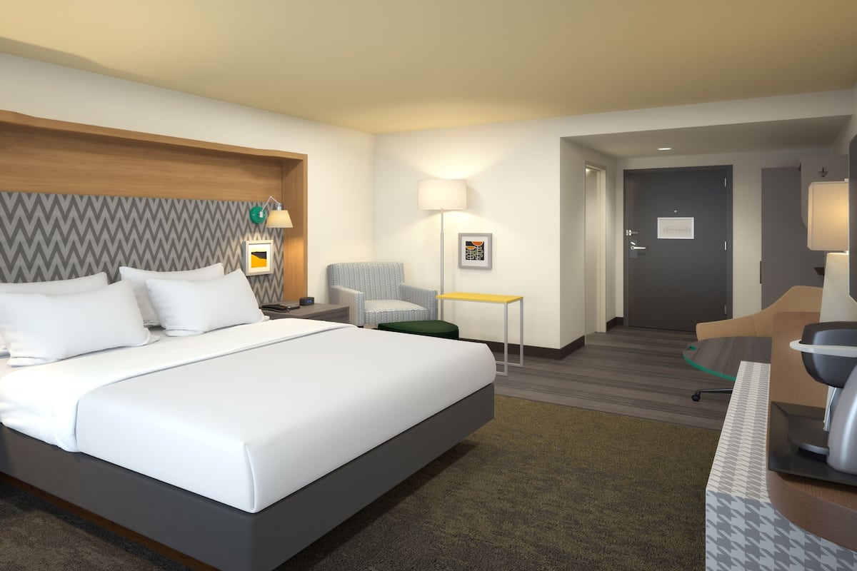 A hotel room with a large bed, a nightstand with a lamp, a desk with a chair, and a door leading to another room.