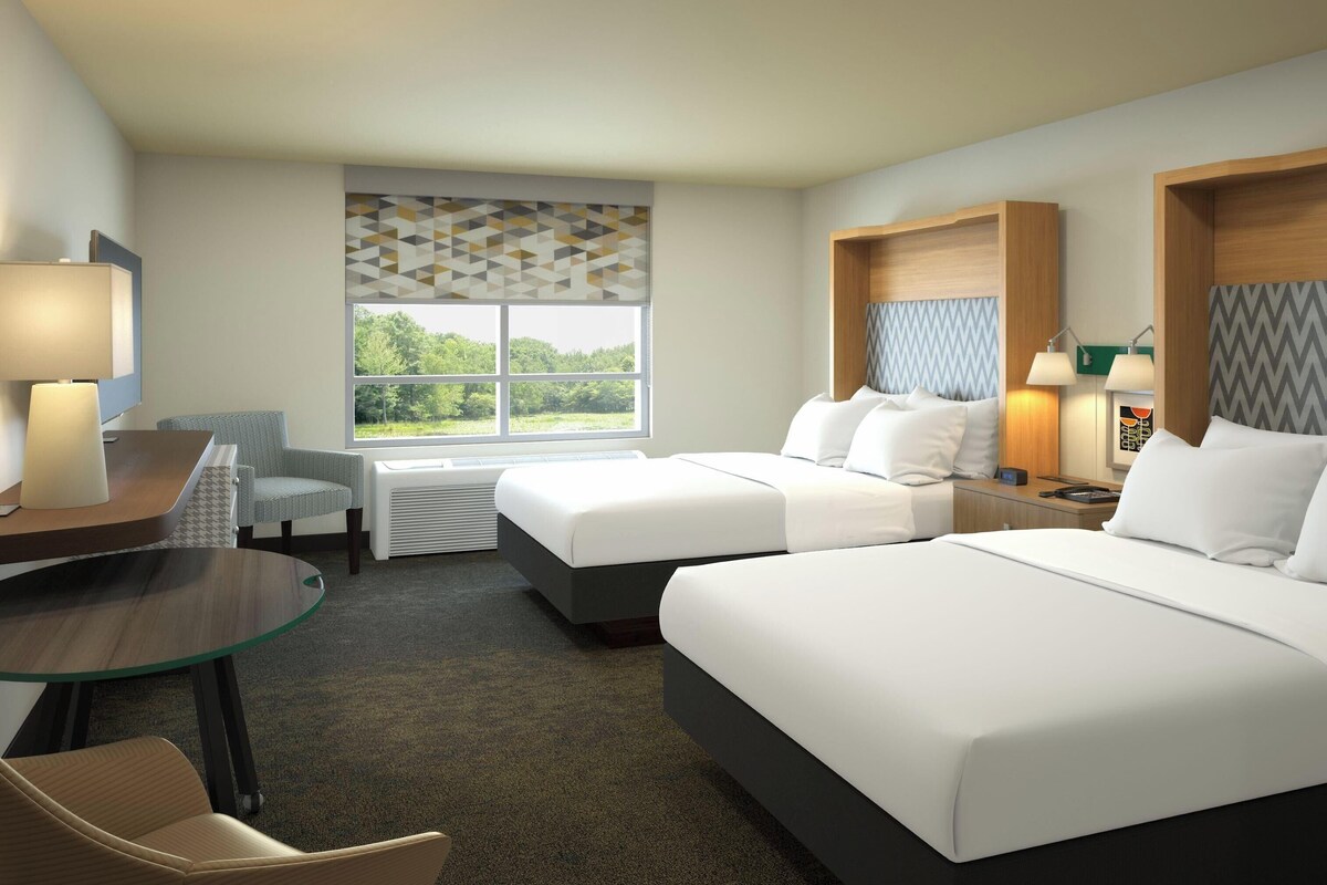 A hotel room with two beds, a desk, a chair, a window with a view, and a heater.