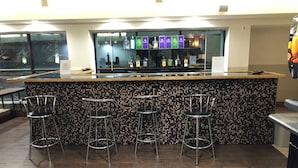 Un espace bar avec un mur recouvert de carreaux, quatre tabourets de bar en métal et une variété de bouteilles d’alcool exposées derrière le comptoir.