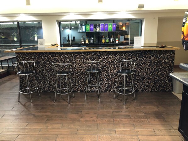 A bar area with a tiled backsplash, four metal bar stools, and a variety of liquor bottles displayed behind the counter. - Holiday Inn Indianapolis Airport Area N by IHG (Indianapolis)