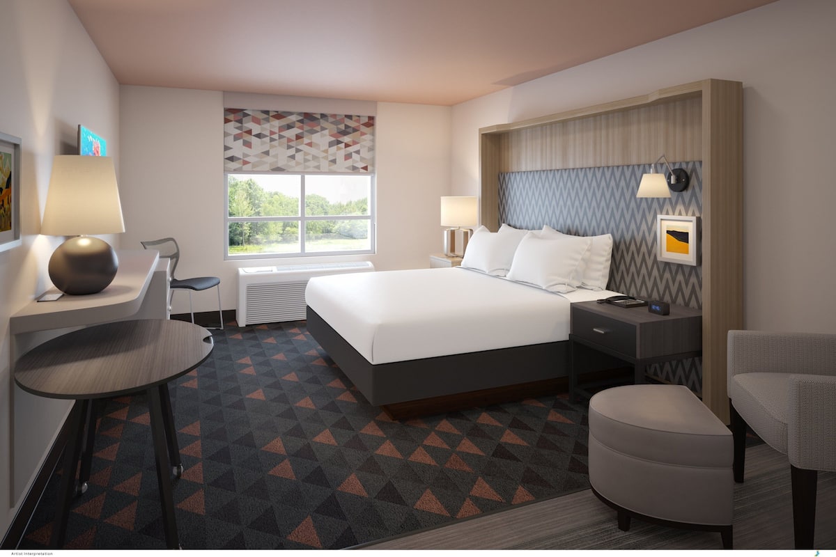 A modern hotel room with a large bed, a desk, a chair, a small table, and a window with a view of greenery.