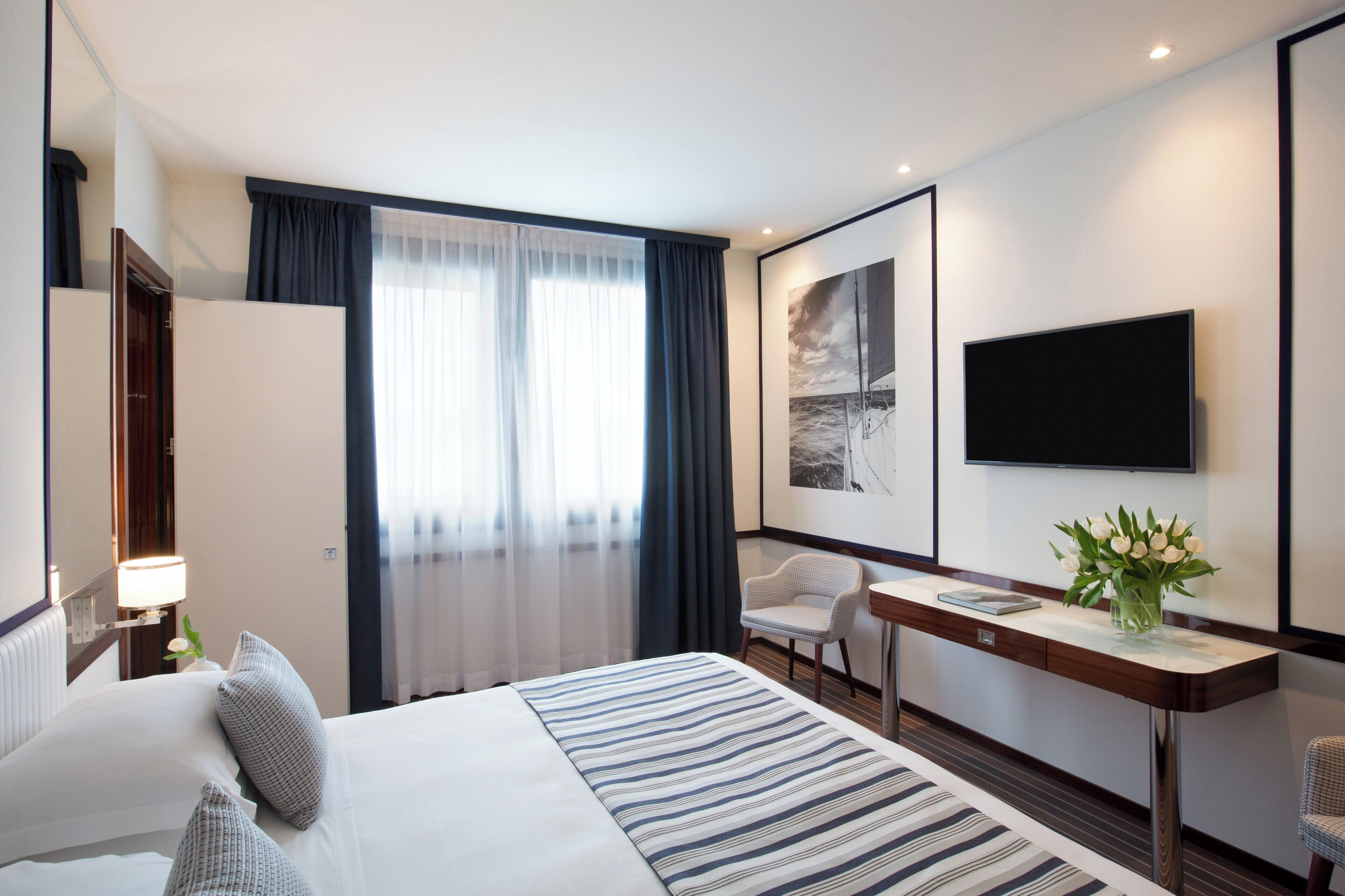 A hotel room with a large bed, a desk with a chair, a flat-screen TV, and a window with curtains.