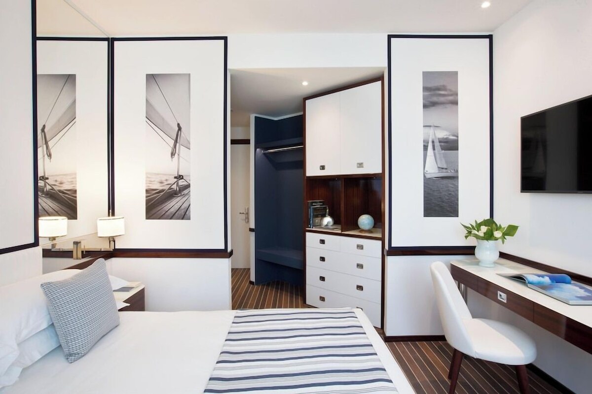 A hotel room with a bed, a desk, a chair, a wardrobe, and framed pictures on the walls.