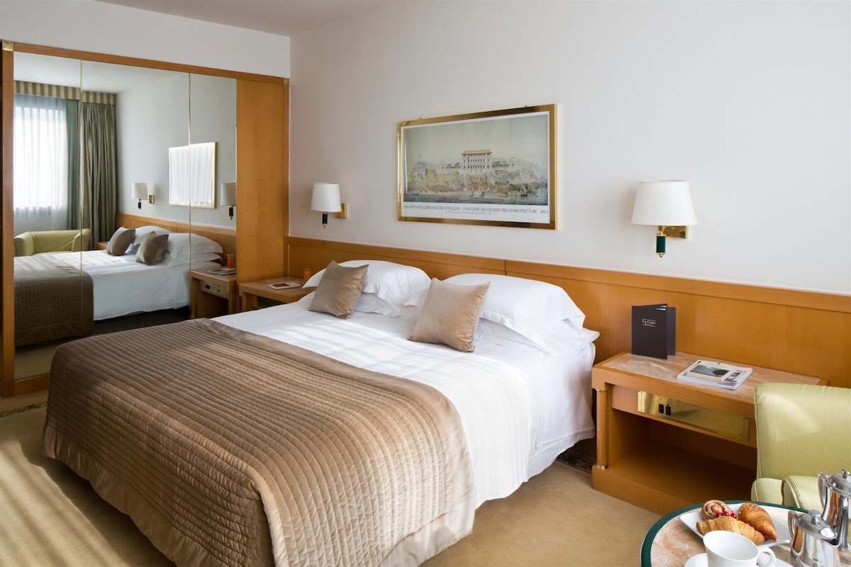 A hotel room with a large bed, two bedside tables, a nightstand, a chair, a table with breakfast items, and a framed picture on the wall.