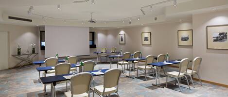 A conference room with tables and chairs, a projector screen, and framed pictures on the walls.