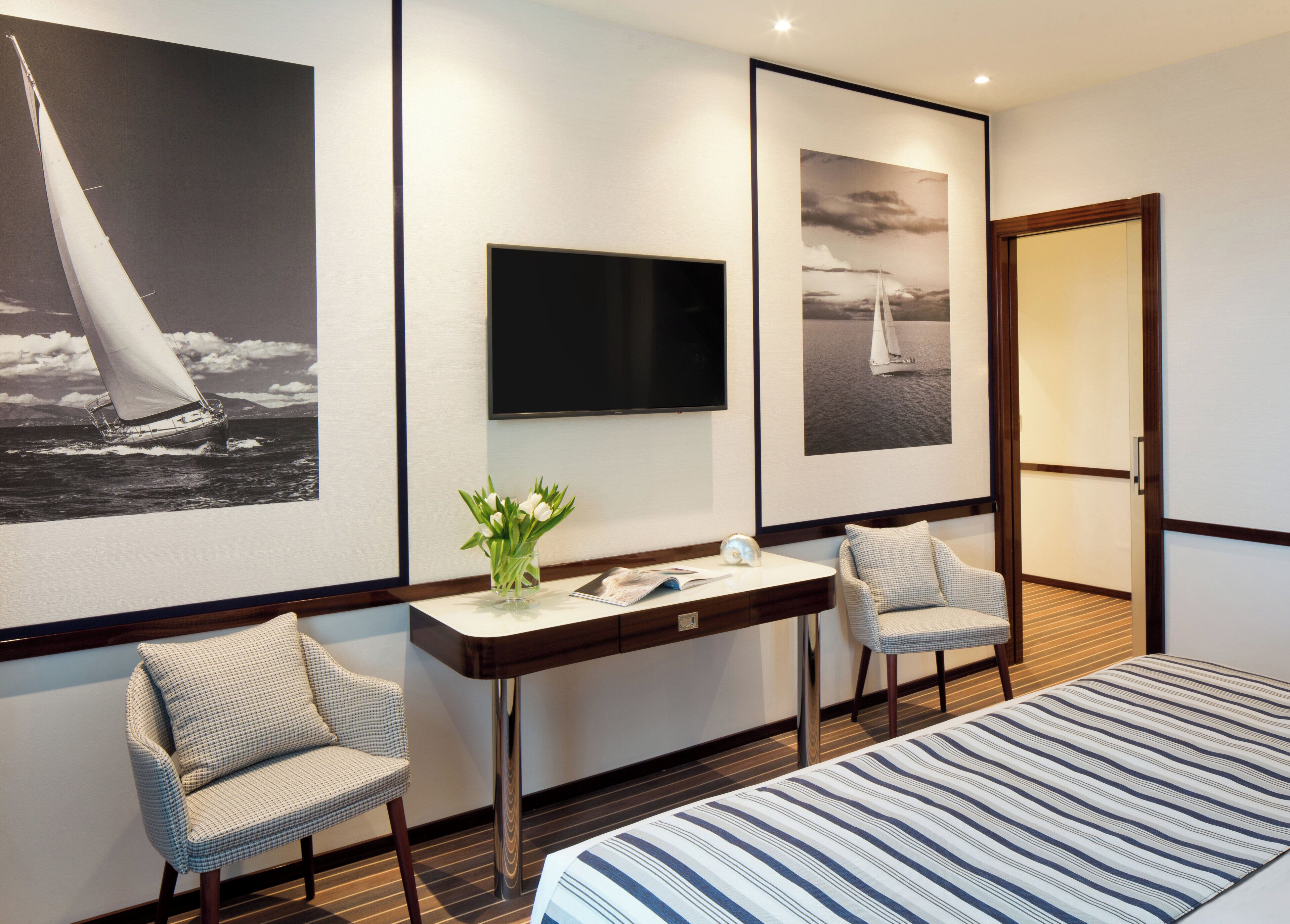 A hotel room with a bed, two chairs, a desk with a flat-screen TV, and two framed pictures of sailboats.